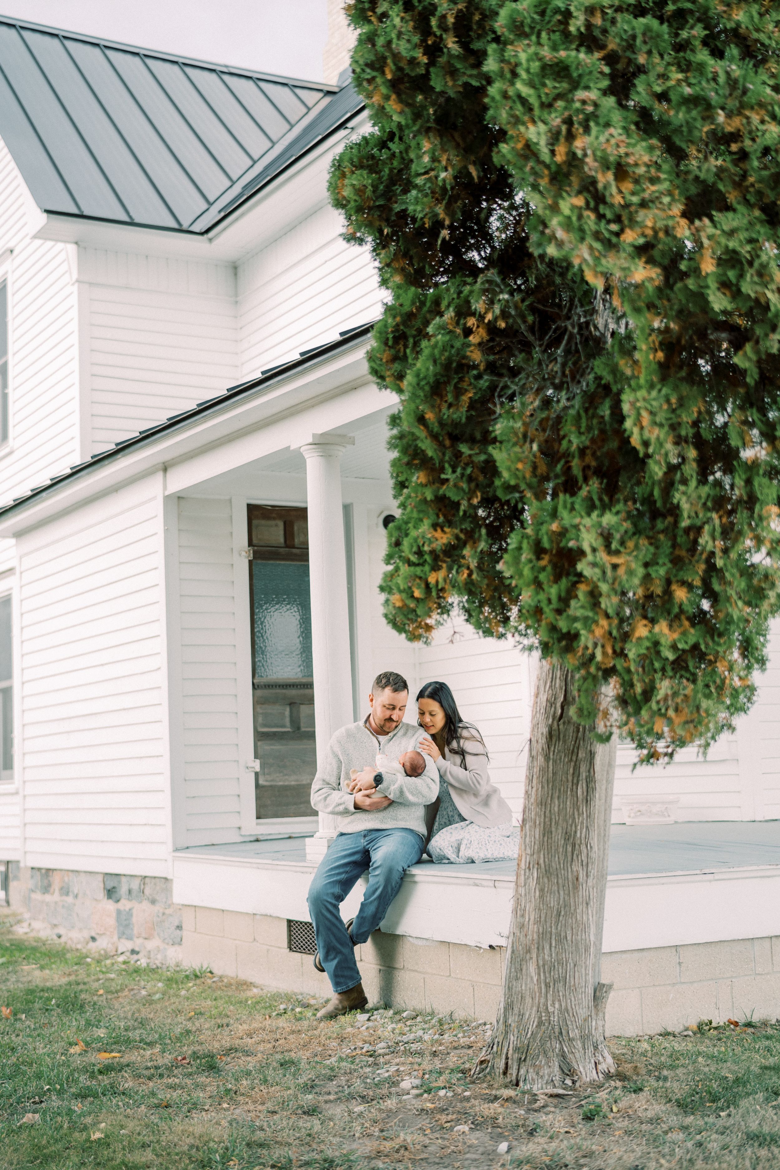 Couple sits together on porch of white farmhouse near large evergreen tree.