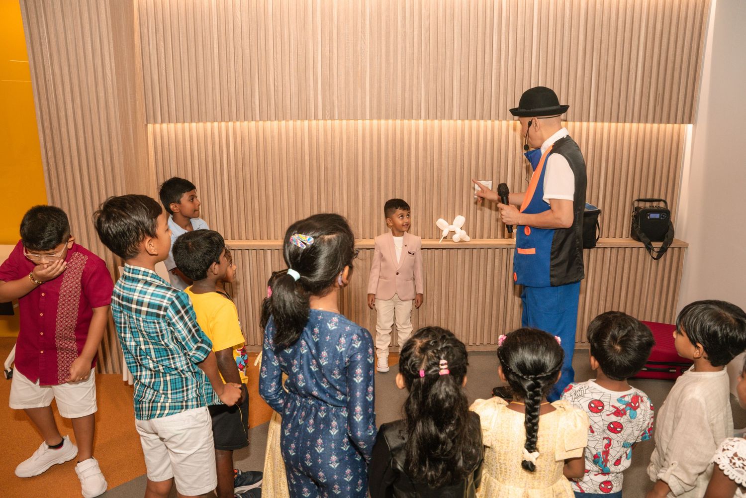 An entertainer in blue overalls and a hat performs for a group of children during a party.