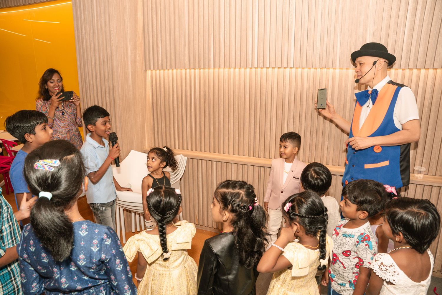 A performer in blue and white holding a microphone entertains a group of engaged children.