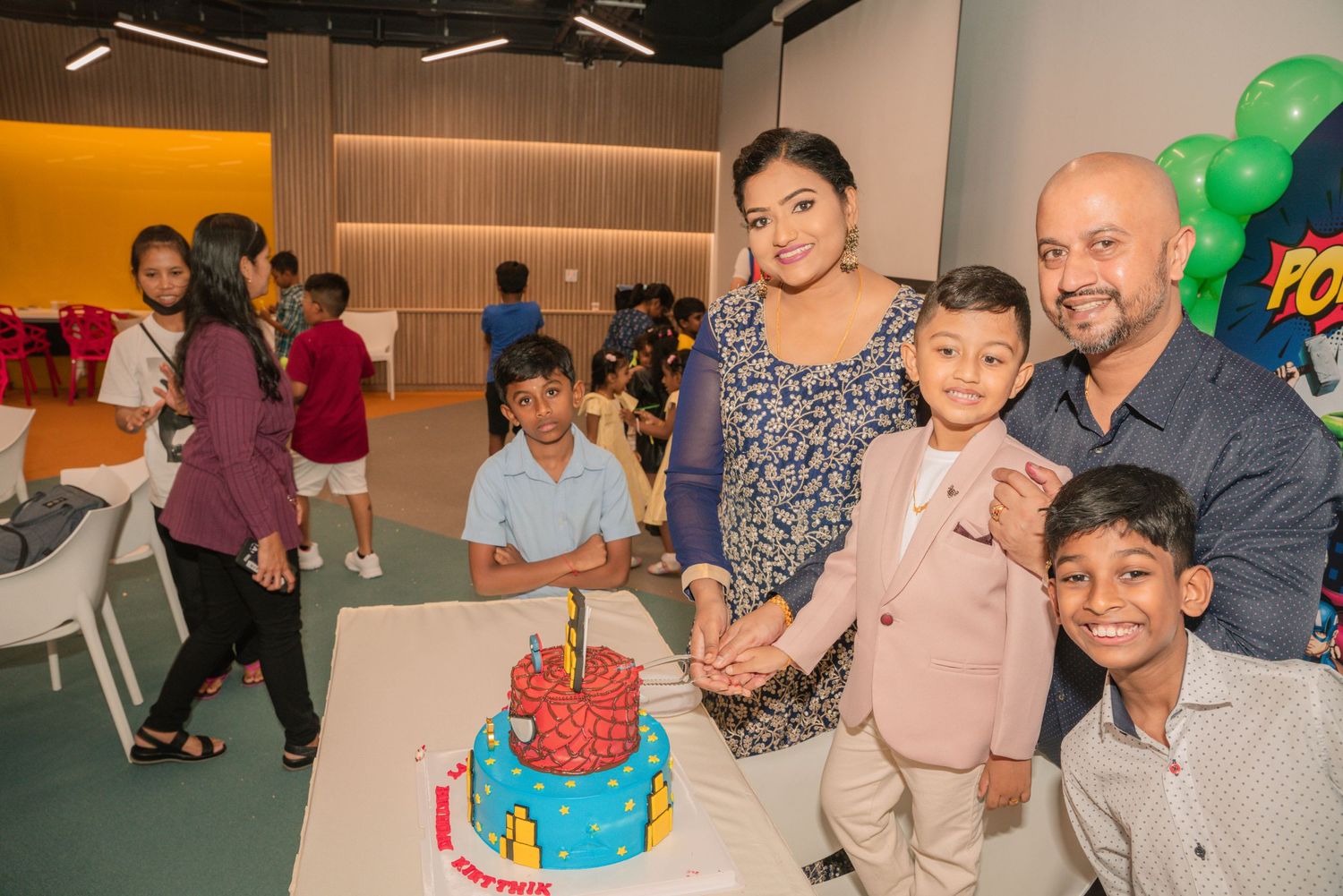 A celebration gathering around a blue Spider-Man themed birthday cake with green balloons in the background.