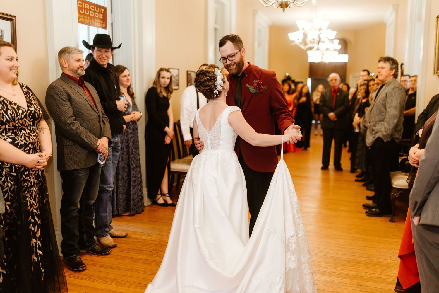 A couple shares their first dance at a wedding reception in an elegant hall with wood floors and onlooking guests.