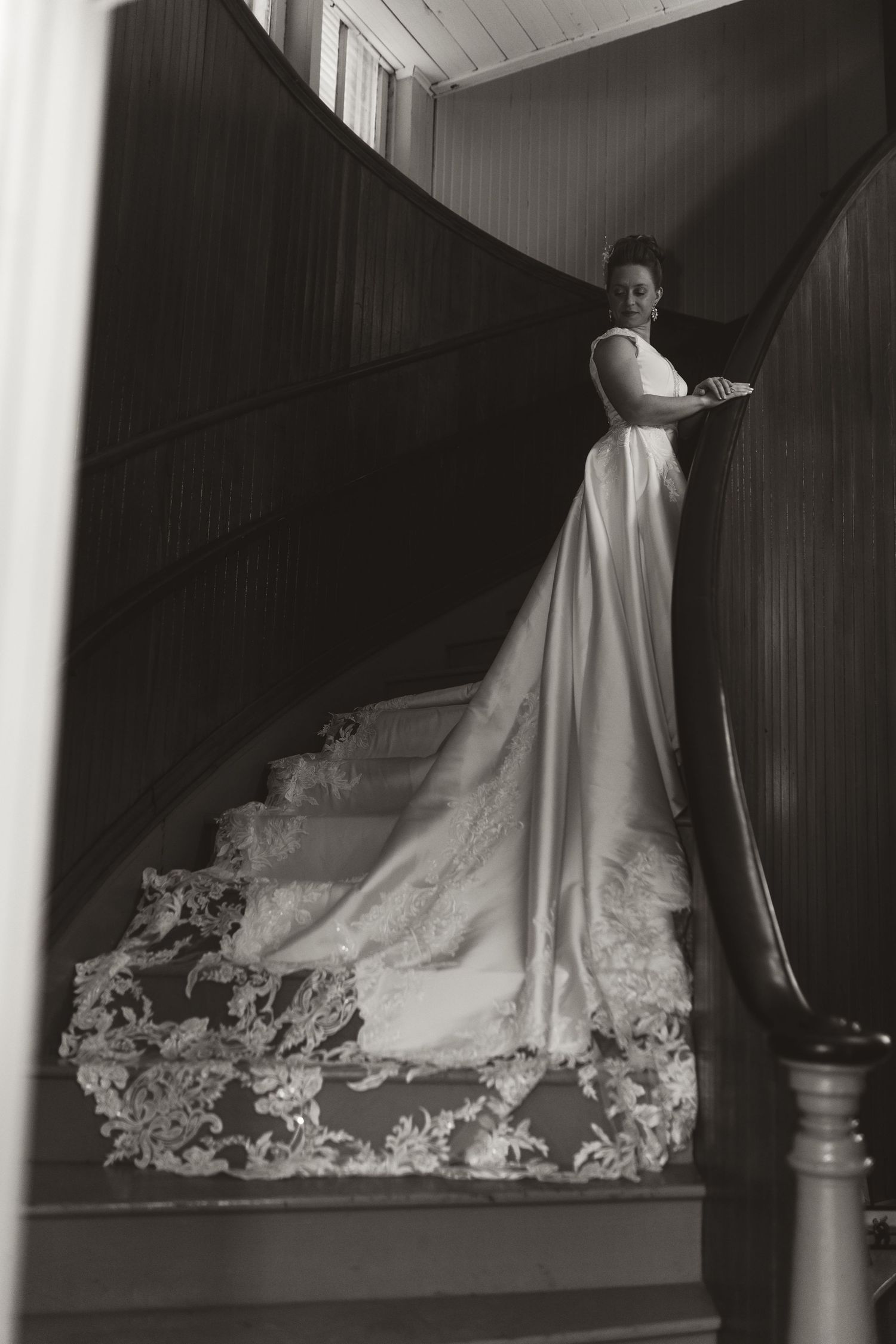 Elegant black and white photograph of figure in flowing wedding gown with ornate lace train on wooden staircase.