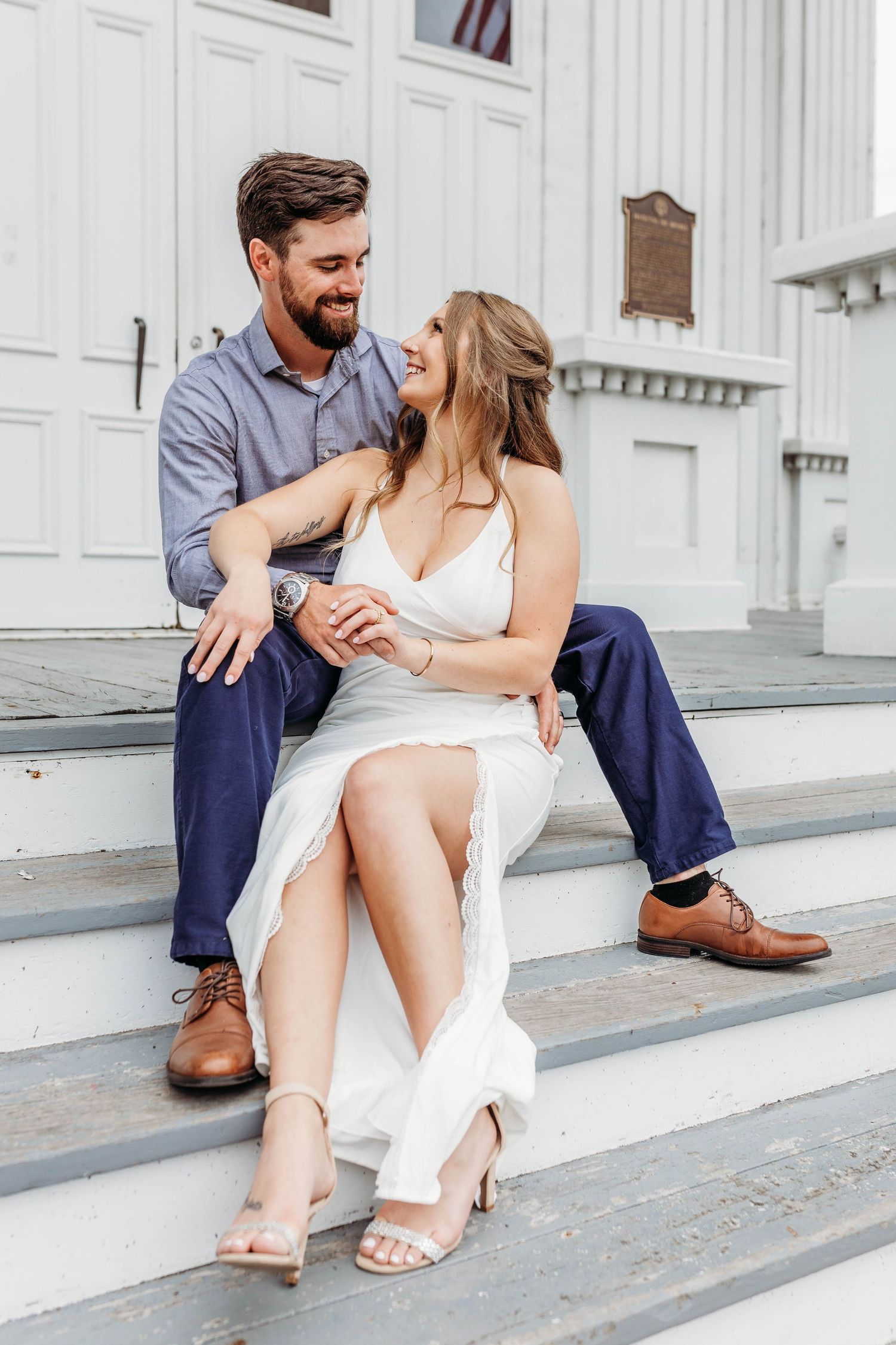 A romantic couple sits together on white outdoor steps, with her in a flowing white dress and him in business casual attire.