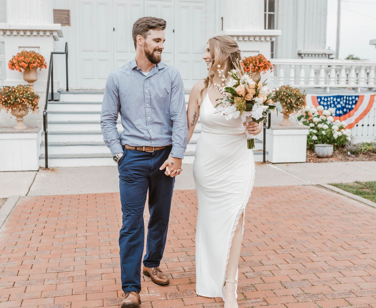 A couple walks together on a brick path next to a white building with floral decorations on a bright day.