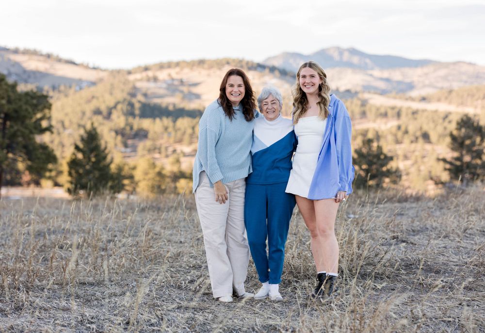 Three people stand together in matching blue tones against a scenic mountain backdrop.