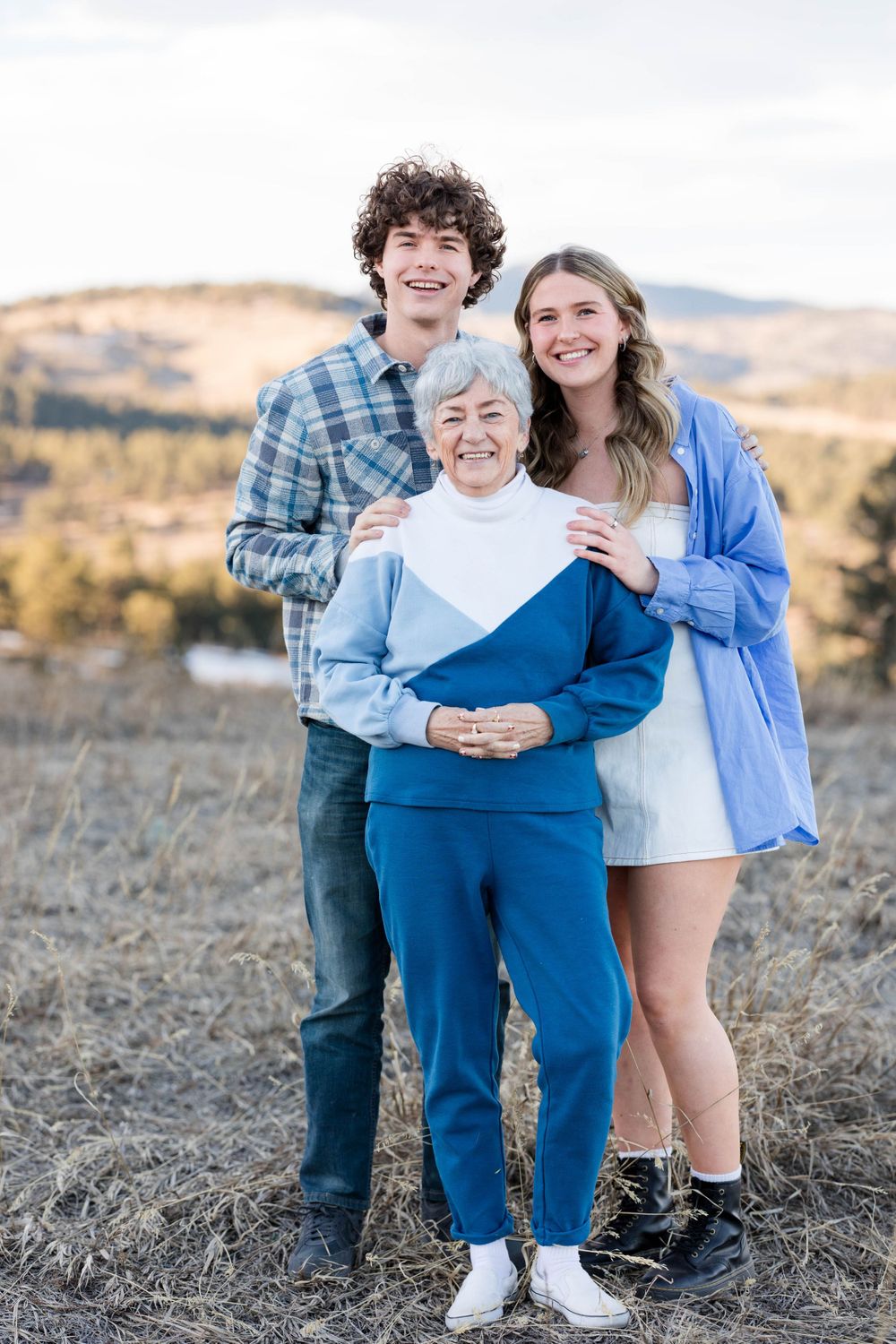 Three people stand close together outdoors with rolling hills in the background.