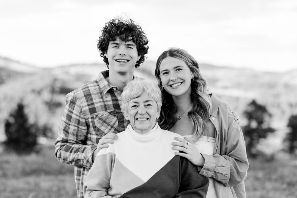 Black and white portrait of three people smiling together with mountains in the distance.