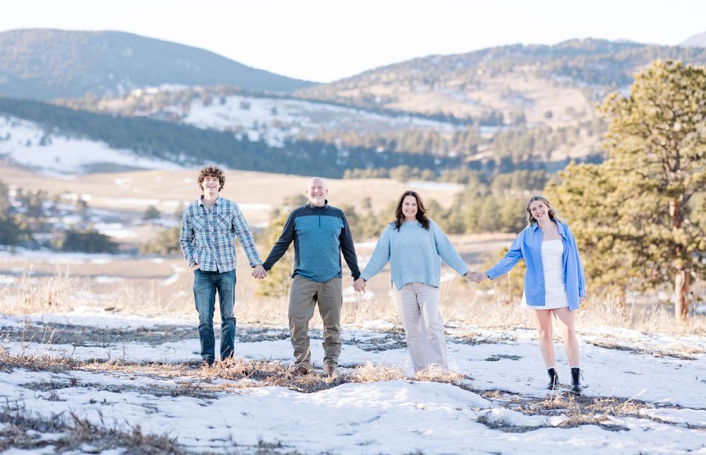 Four people hold hands while walking along a snowy mountain path with scenic mountain views.