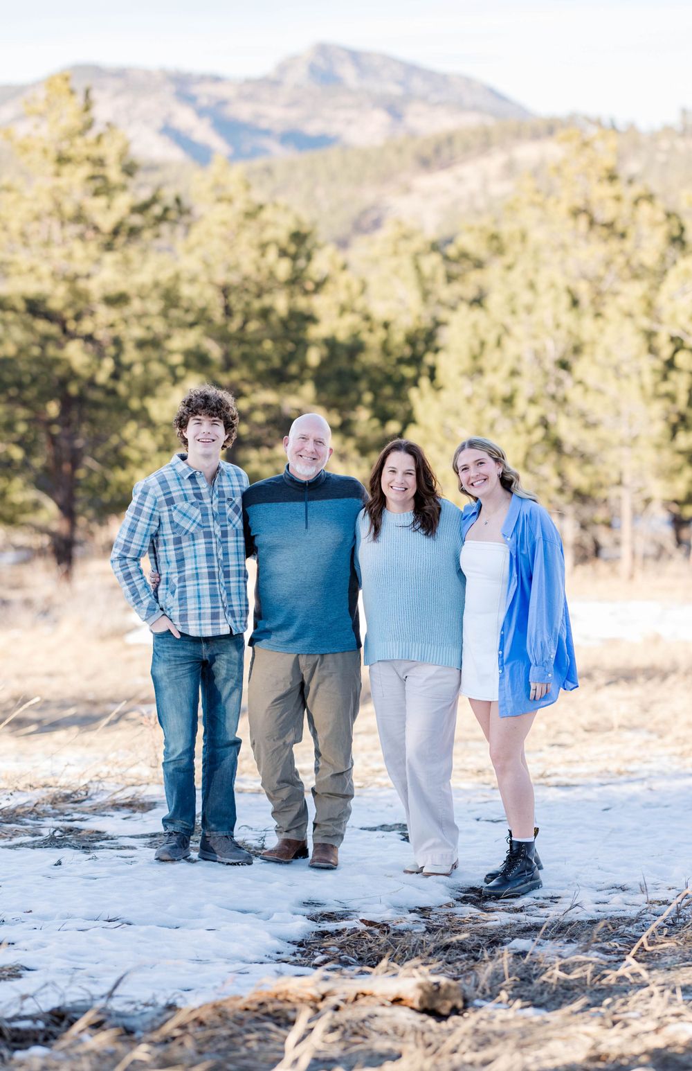 A family stands together on a snowy mountain trail with pine trees and mountains in the background.