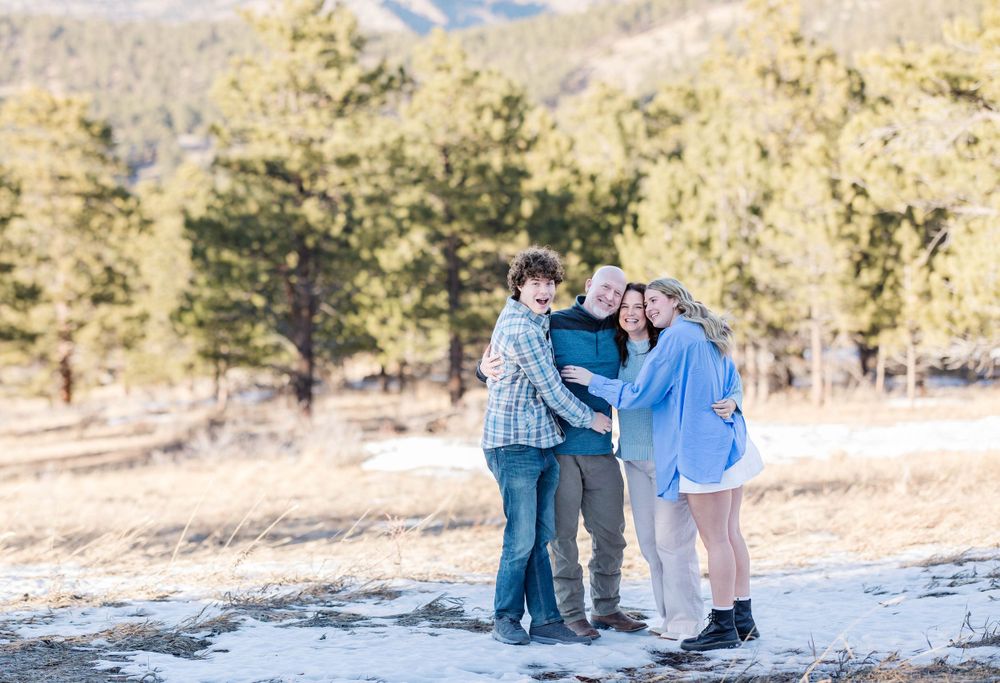 A group embraces while standing together on a snowy mountain path with evergreen trees in the background.