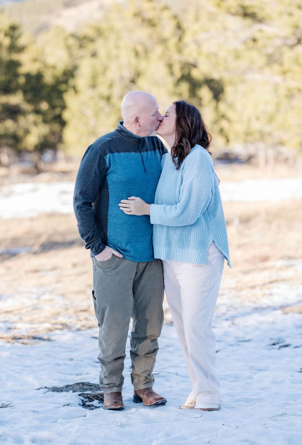 A couple shares an intimate moment while standing on snow-covered ground with mountains behind them.