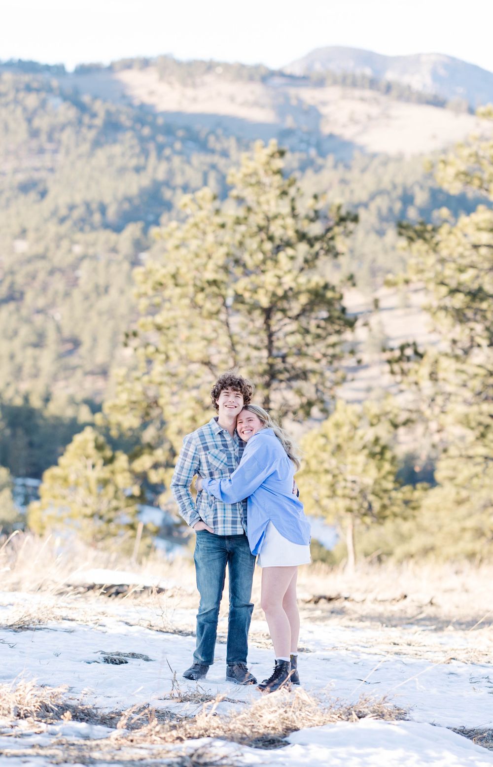 A couple stands close together on a rocky mountain overlook with scenic forest views behind them.