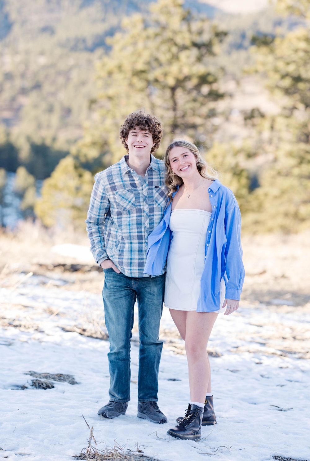 A couple poses together outdoors on rocky terrain with mountain scenery and pine trees in the background.
