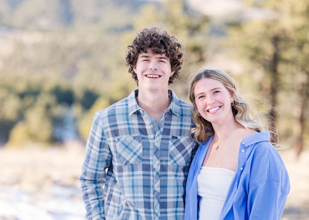 A couple smiles at the camera while standing outdoors with mountains and forest in the distance.