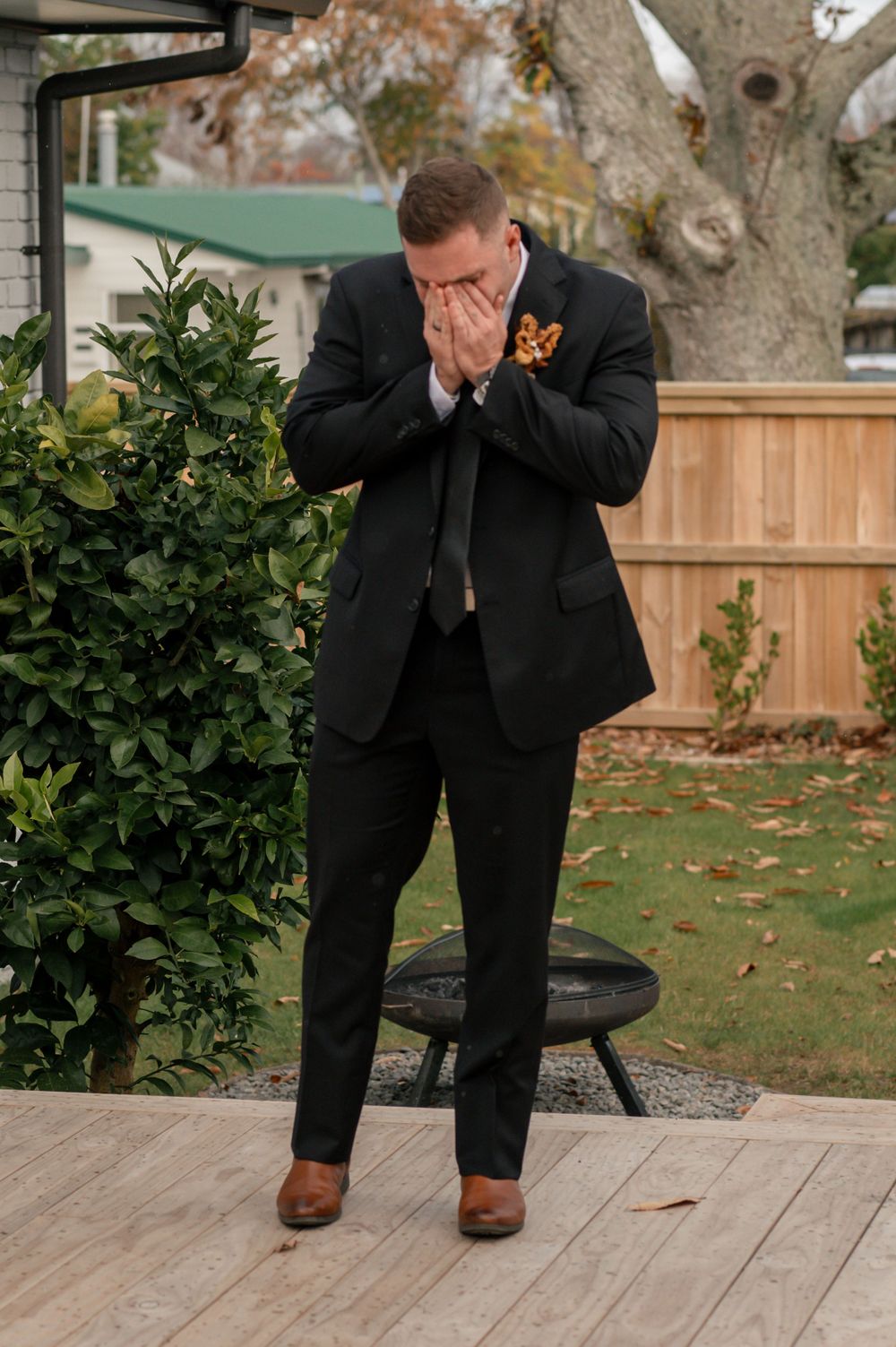 Person in black suit with orange boutonniere shows emotional reaction on outdoor wooden deck Cambridge New Zealand