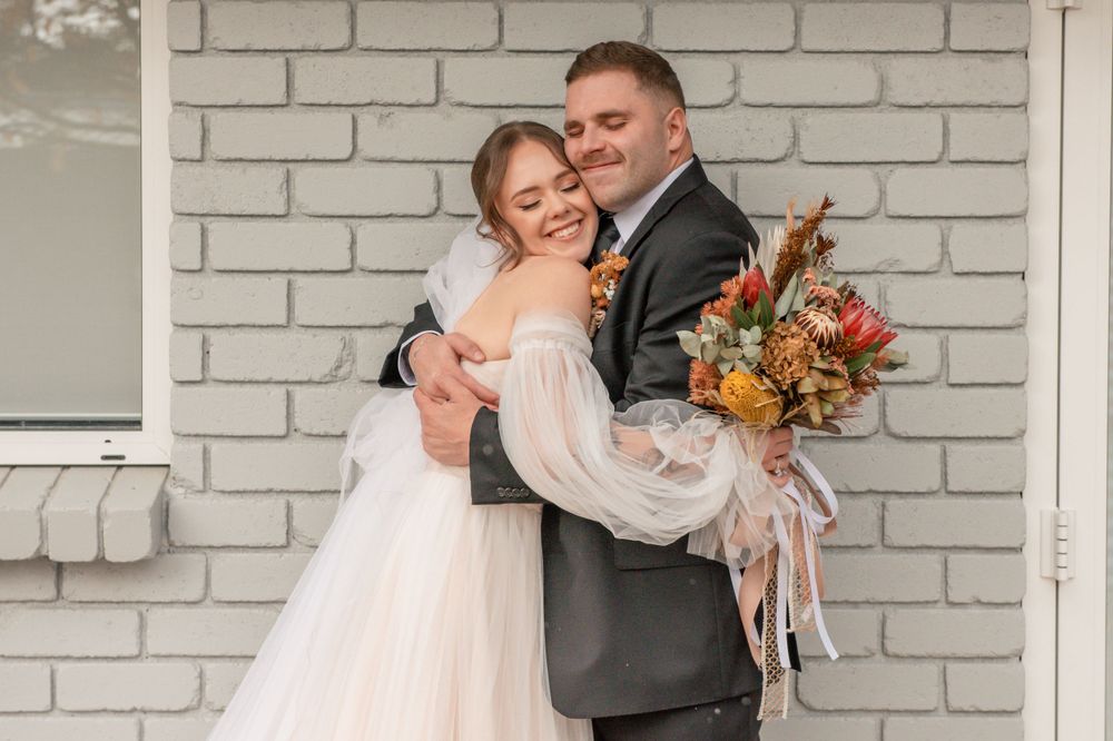 Joyful wedding couple poses against white brick wall with fall-colored bouquet Cambridge New Zealand