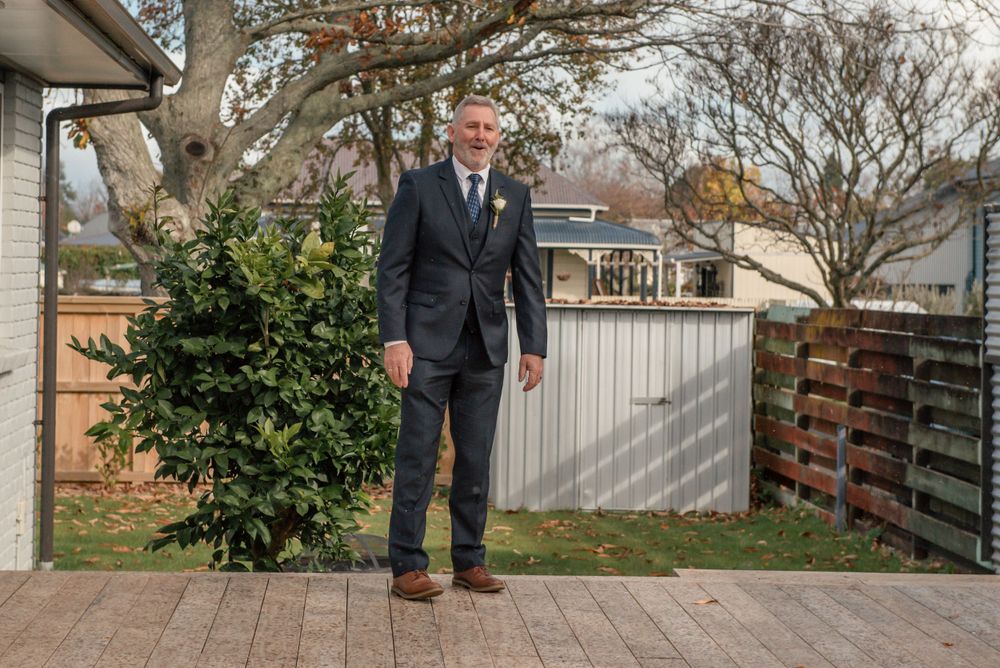 Person in navy suit stands on wooden deck near green shrub and white fence. Cambridge New Zealand
