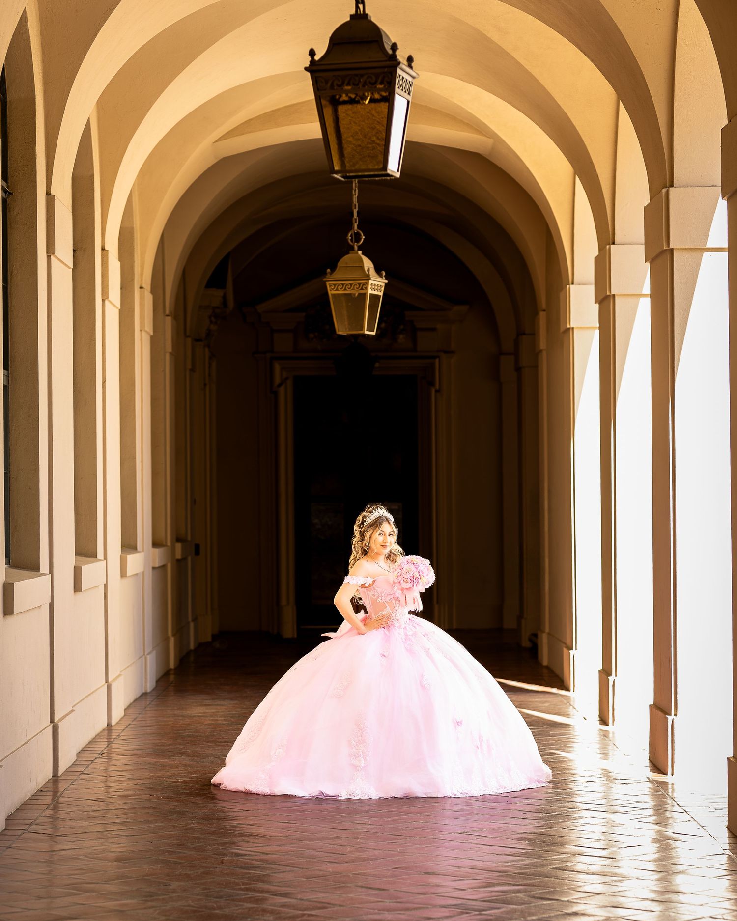 Quinceañera poses in a stunning pink dress down an elegant hallway. Professional photographer photographs Quinceañera using natural lighting