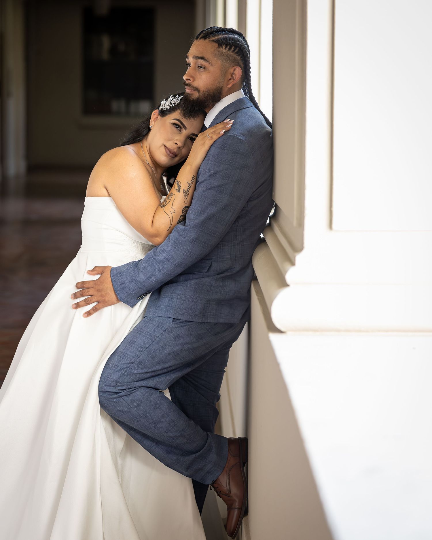 A romantic wedding moment captured as a bride and groom embrace against a white column in soft natural lighting.