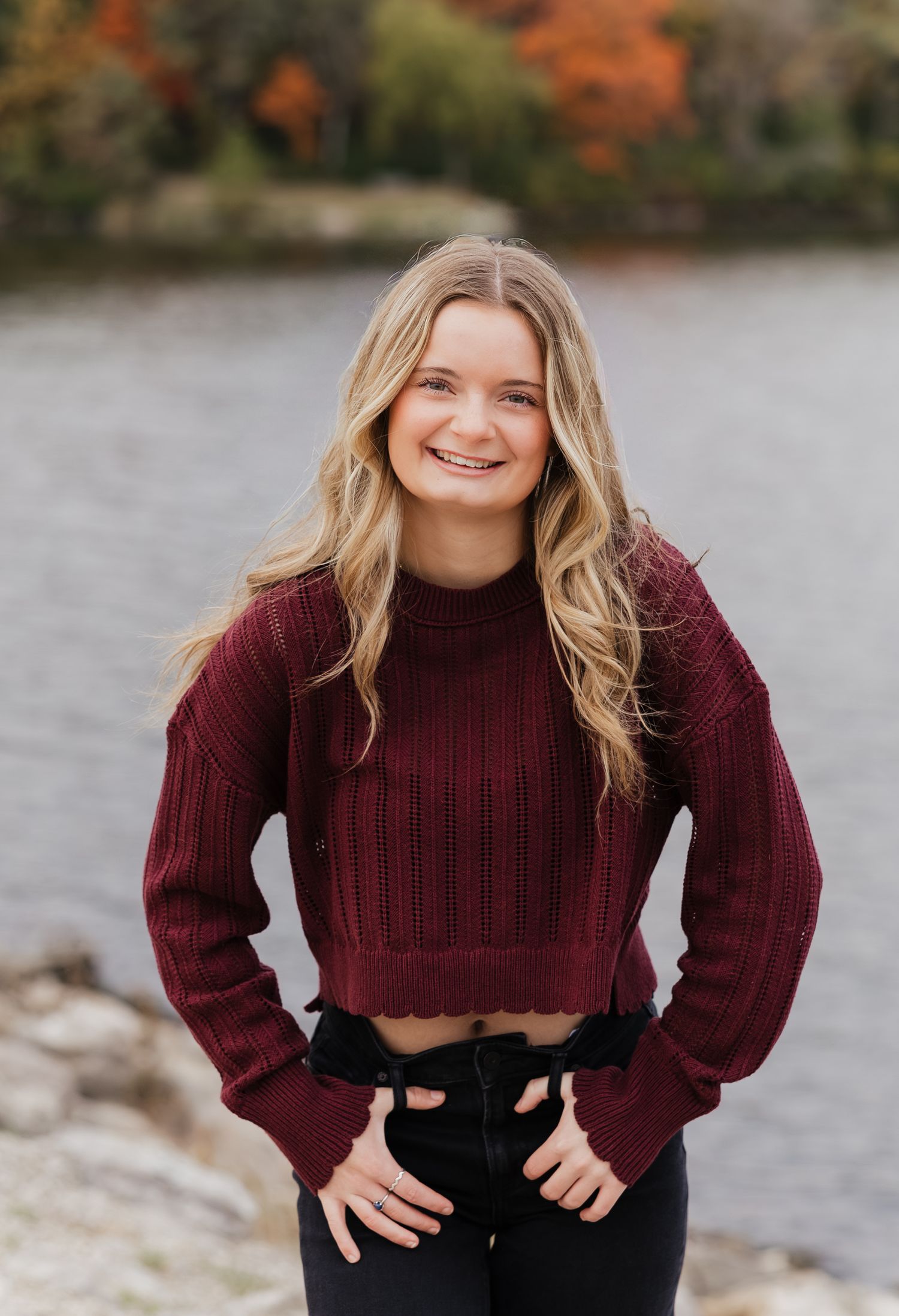 A person wearing a burgundy knit sweater poses against an autumn lakeside backdrop.