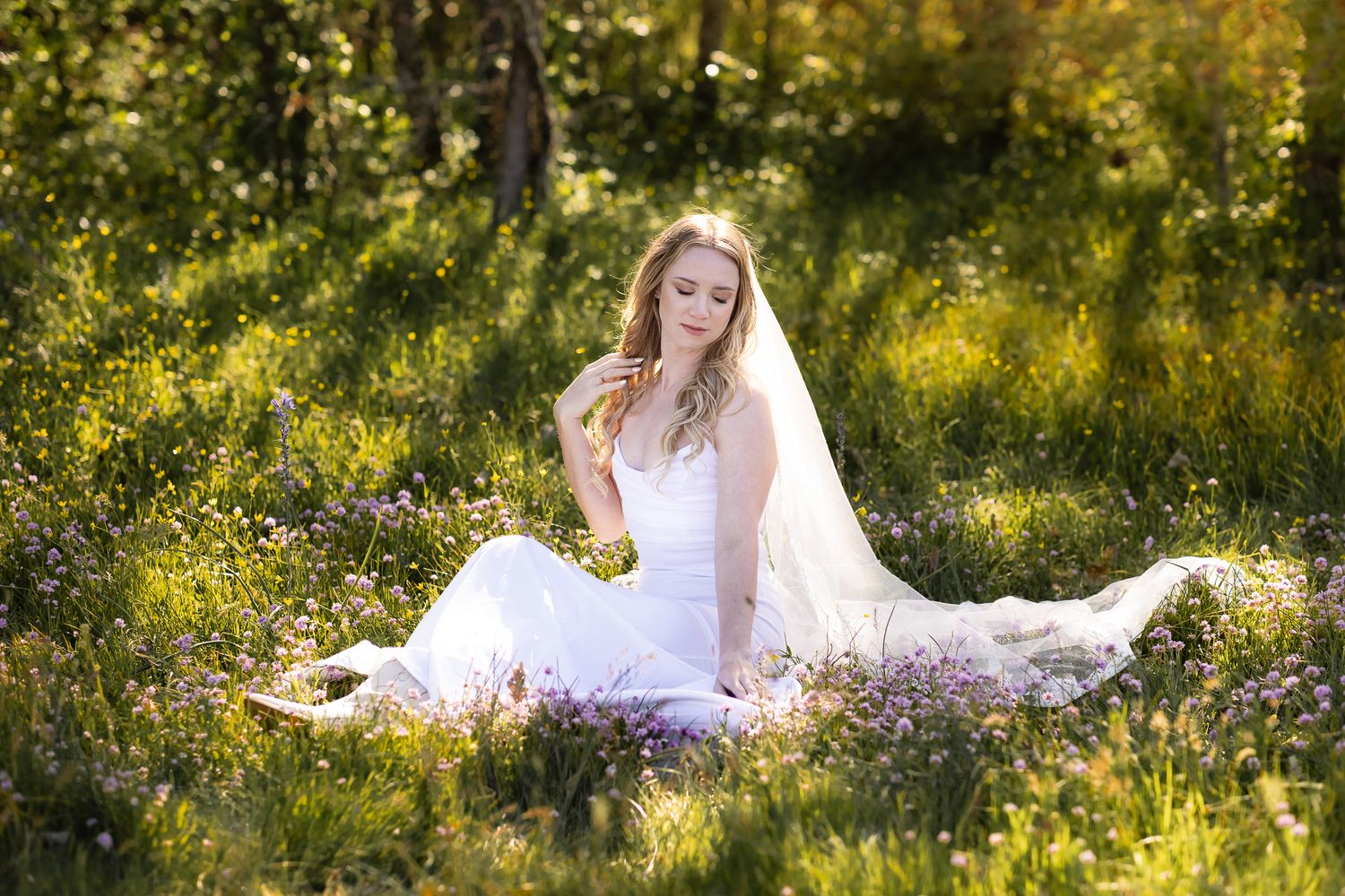 A bride with a veil sits in sun-dappled grass, surrounded by tiny wildflowers in southern Washington.
