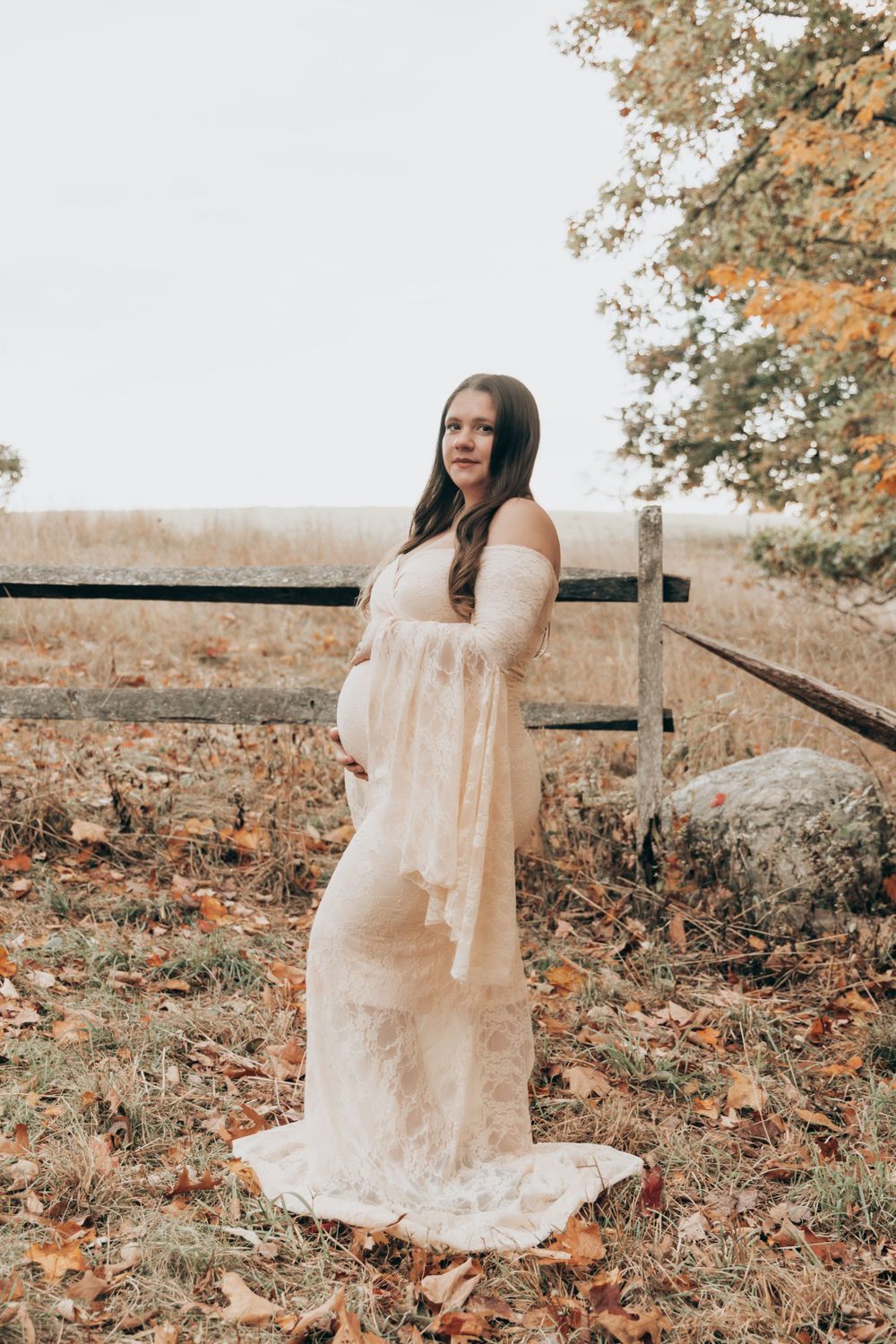 Person in cream lace maternity gown poses in rustic autumn setting with fence and fallen leaves.