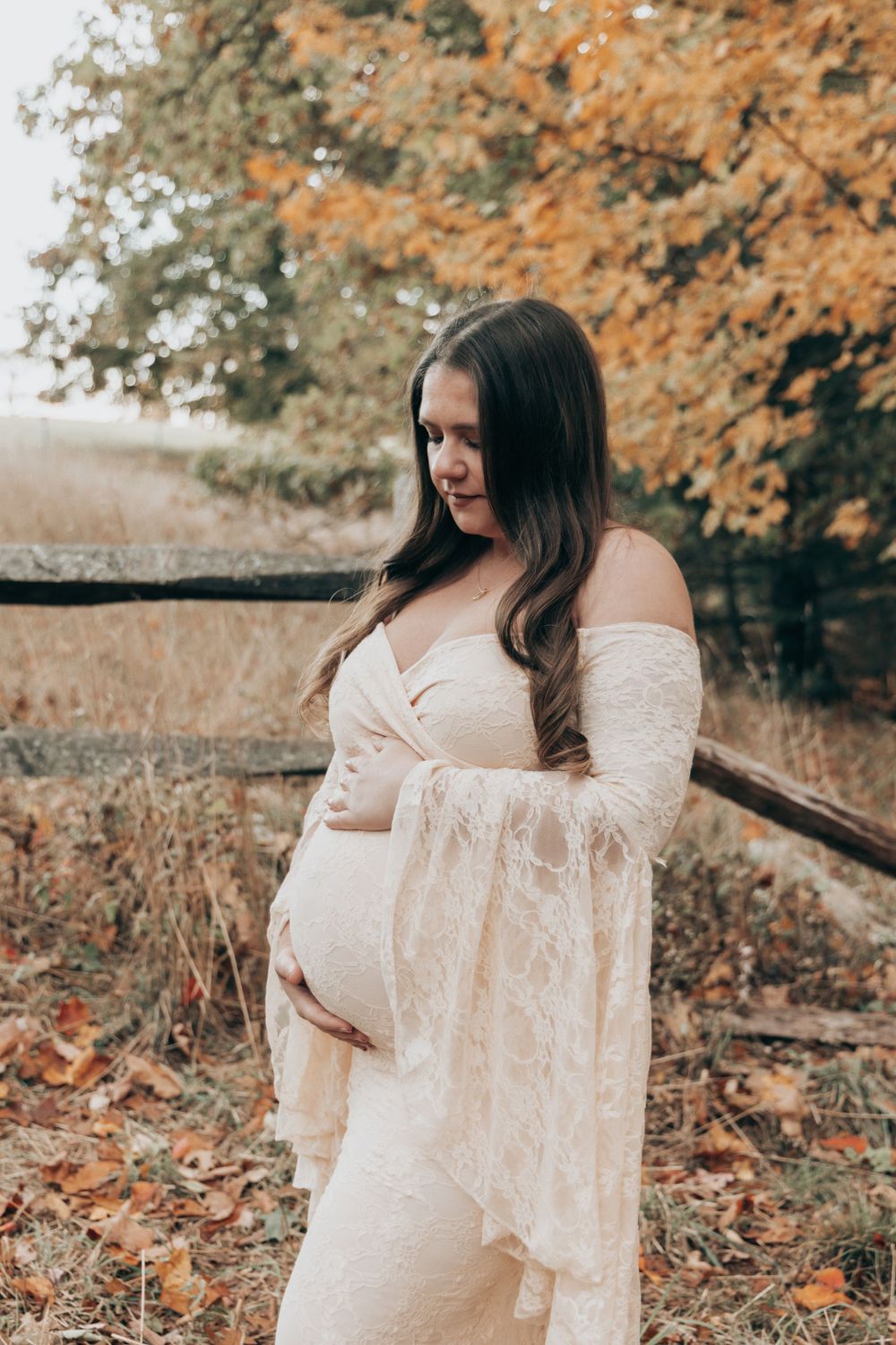 Maternity portrait in white lace dress against autumn foliage backdrop.
