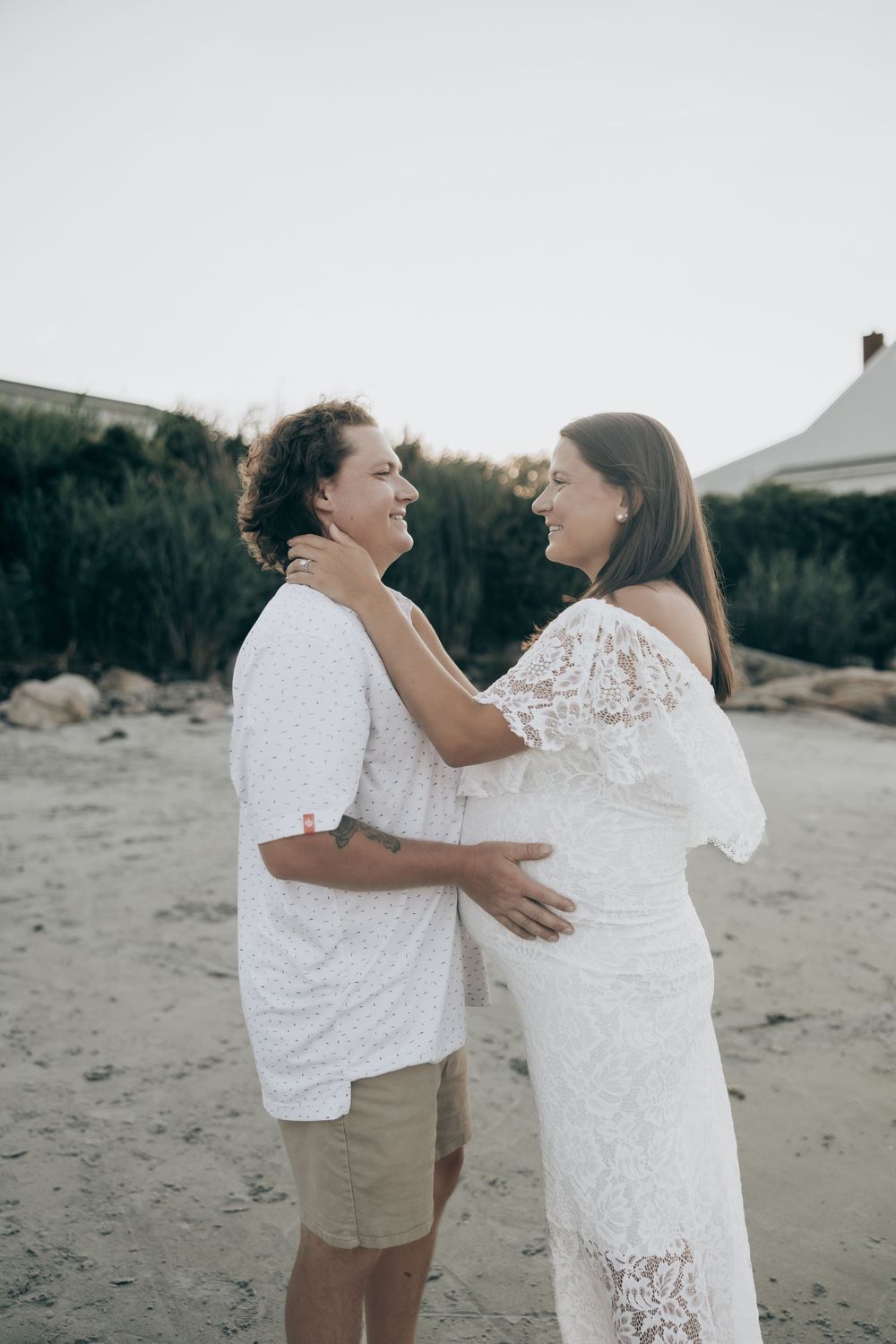 Two people wearing white outfits share a joyful moment laughing together on a sandy beach at sunset.