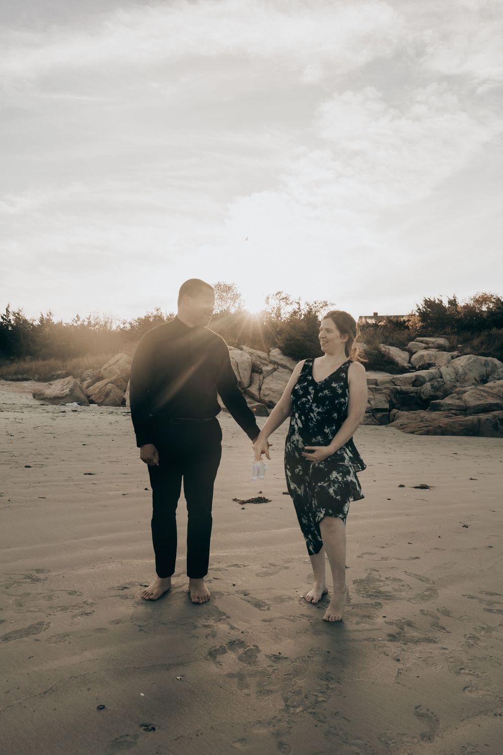 Couple walking hand in hand on a beach at sunset with rocky outcrop in background.