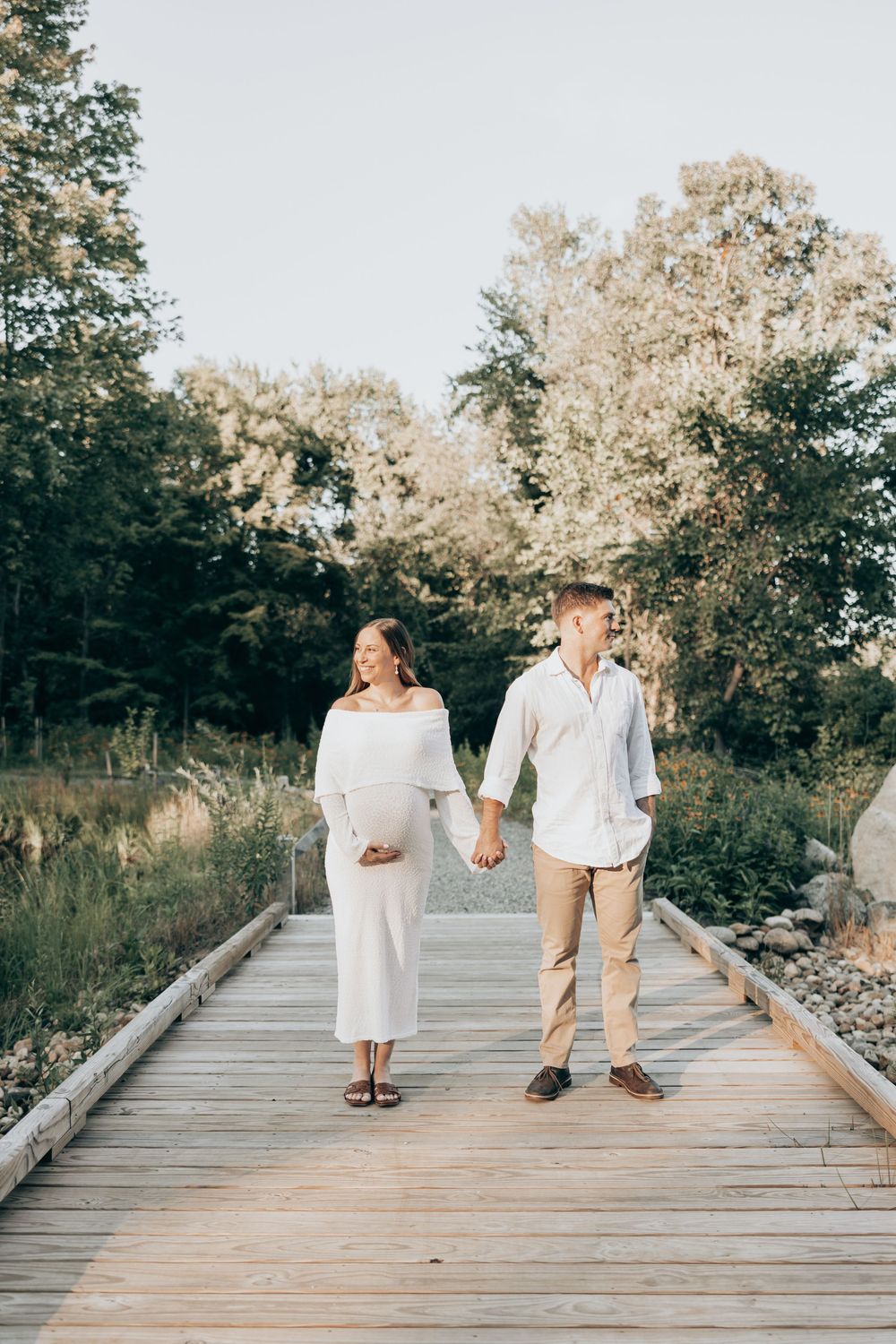 A couple holds hands while walking on a wooden boardwalk surrounded by lush greenery on a sunny day.