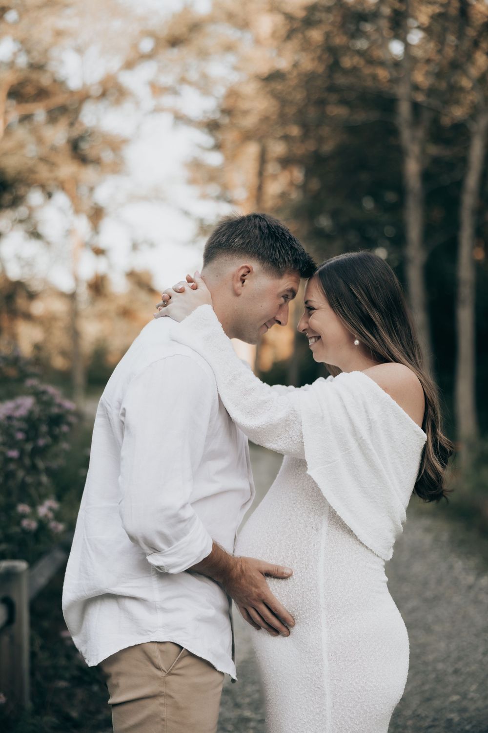A romantic maternity photo of a couple in white outfits sharing an intimate moment in a wooded outdoor setting.