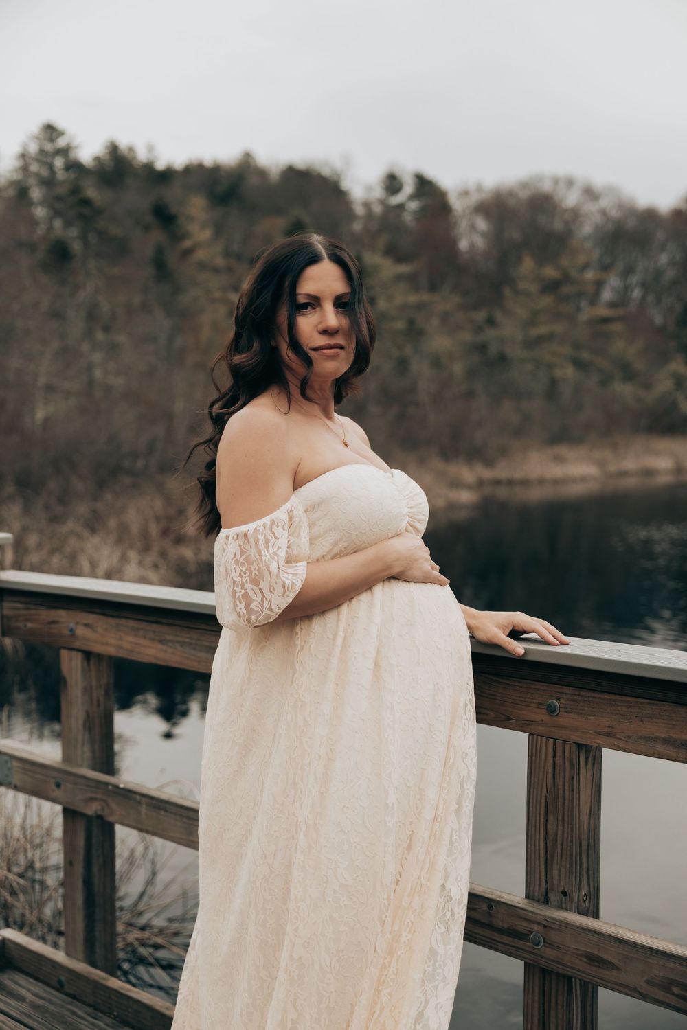 Expecting mother in white lace dress poses on wooden bridge over calm lake during maternity photoshoot.