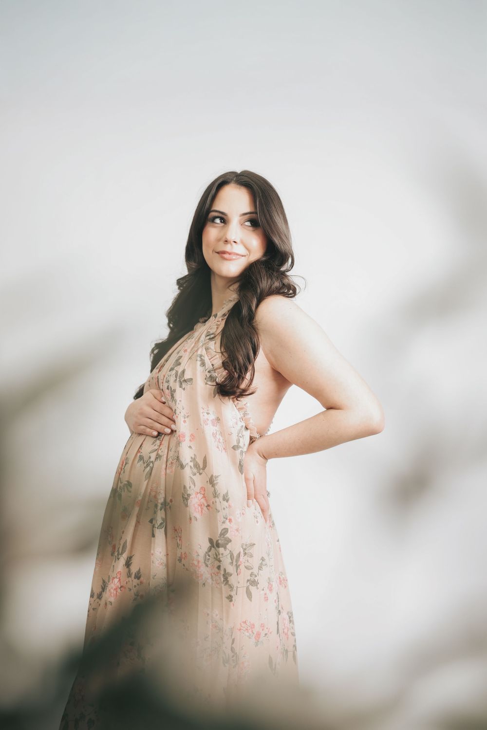 Pregnant woman in floral dress cradling baby bump, smiling softly against light background.