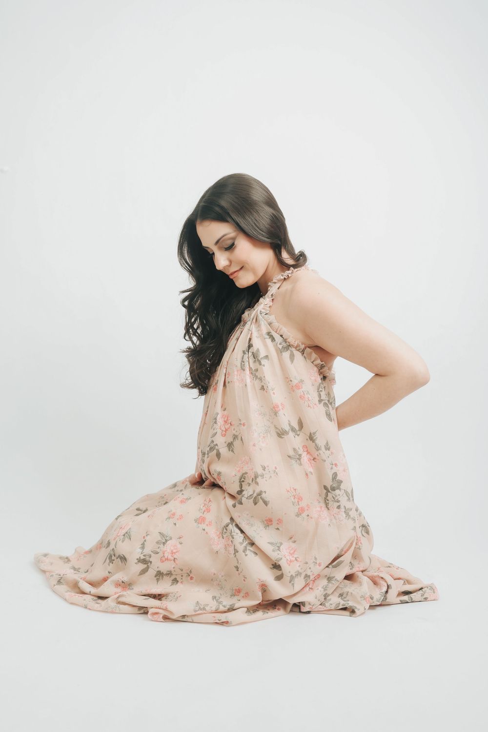 Pregnant woman in floral halter dress sitting gracefully on floor, smiling downward.
