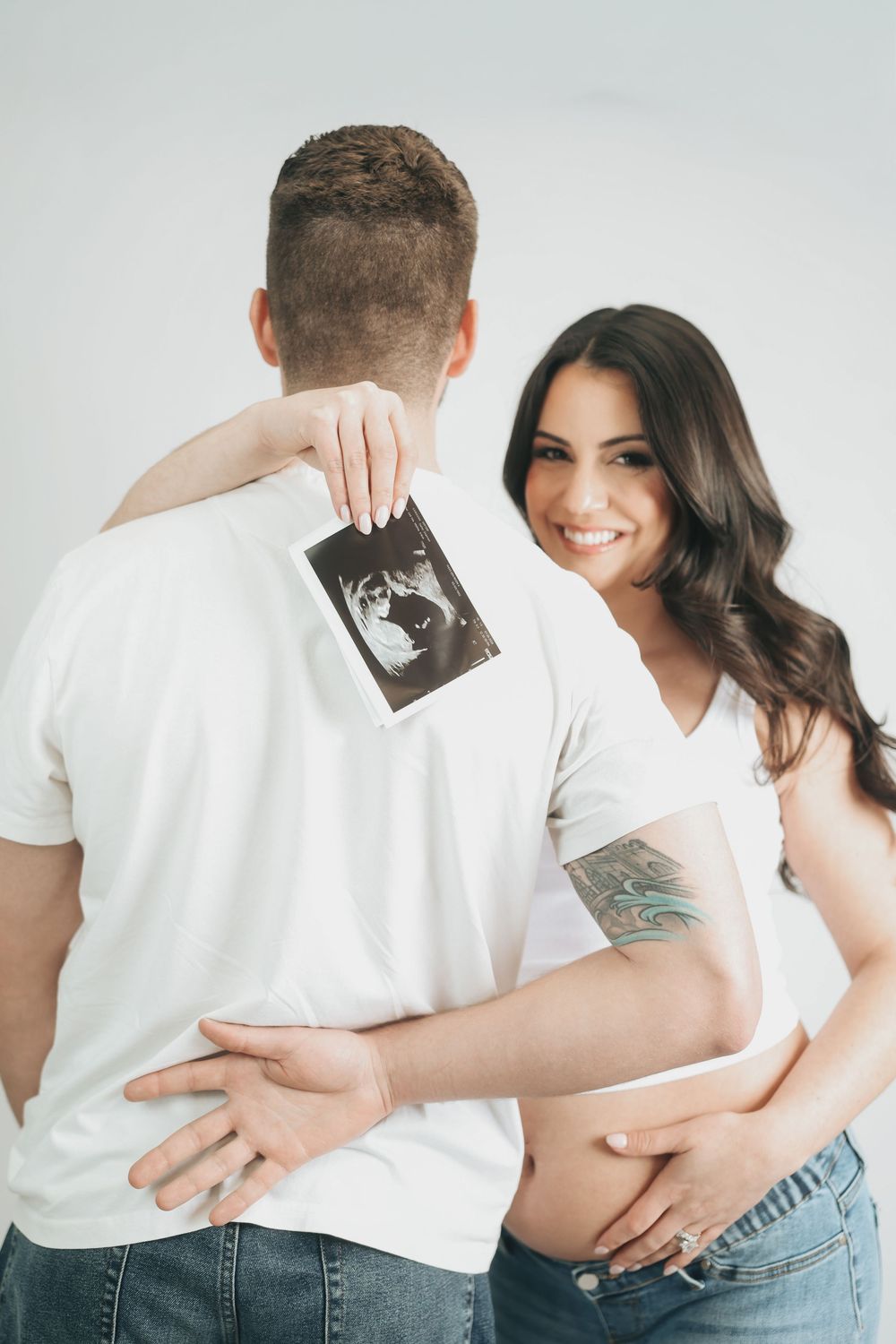 Happy pregnant couple embracing, woman smiling while man holds ultrasound photo behind his back.