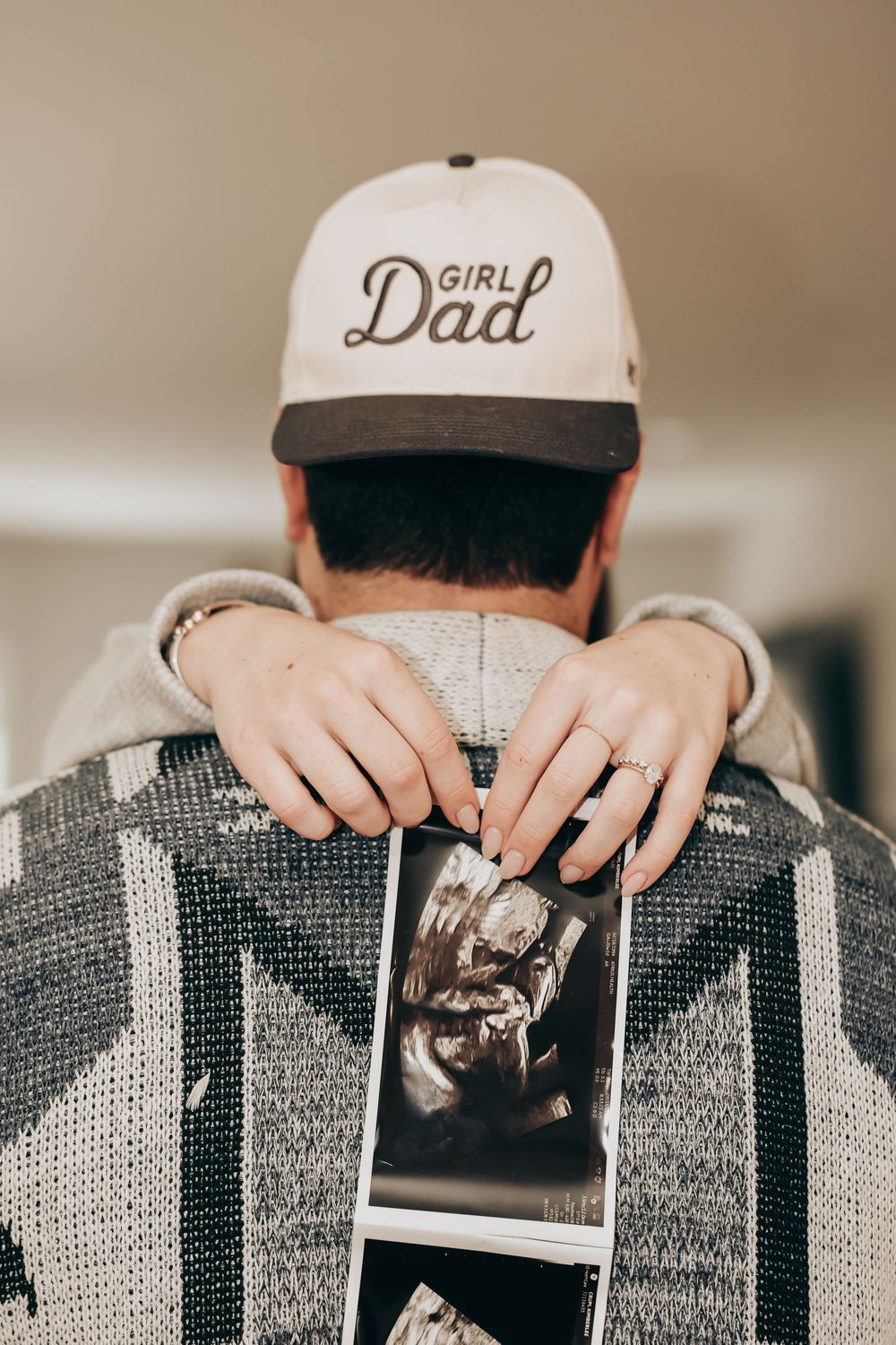 Person wearing a 'Girl Dad' baseball cap holds up an ultrasound photo while wearing a gray patterned sweater.