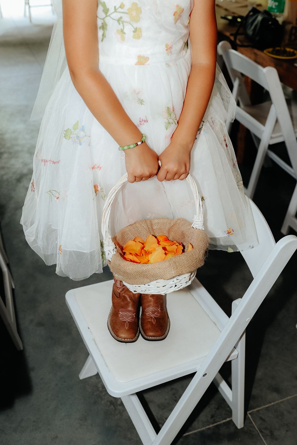 A flower girl in a white dress holds a basket while sitting on white folding chairs.