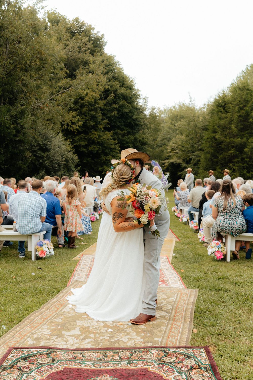 Wedding guests seated on vintage rugs during an outdoor ceremony in a grassy field.