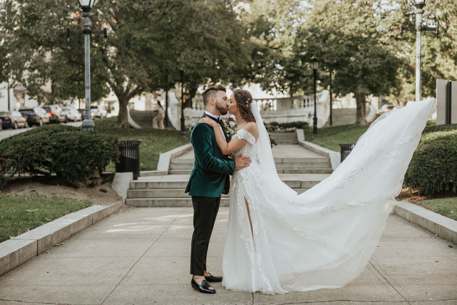 Wedding couple embracing on sidewalk with flowing white veil catching the breeze.