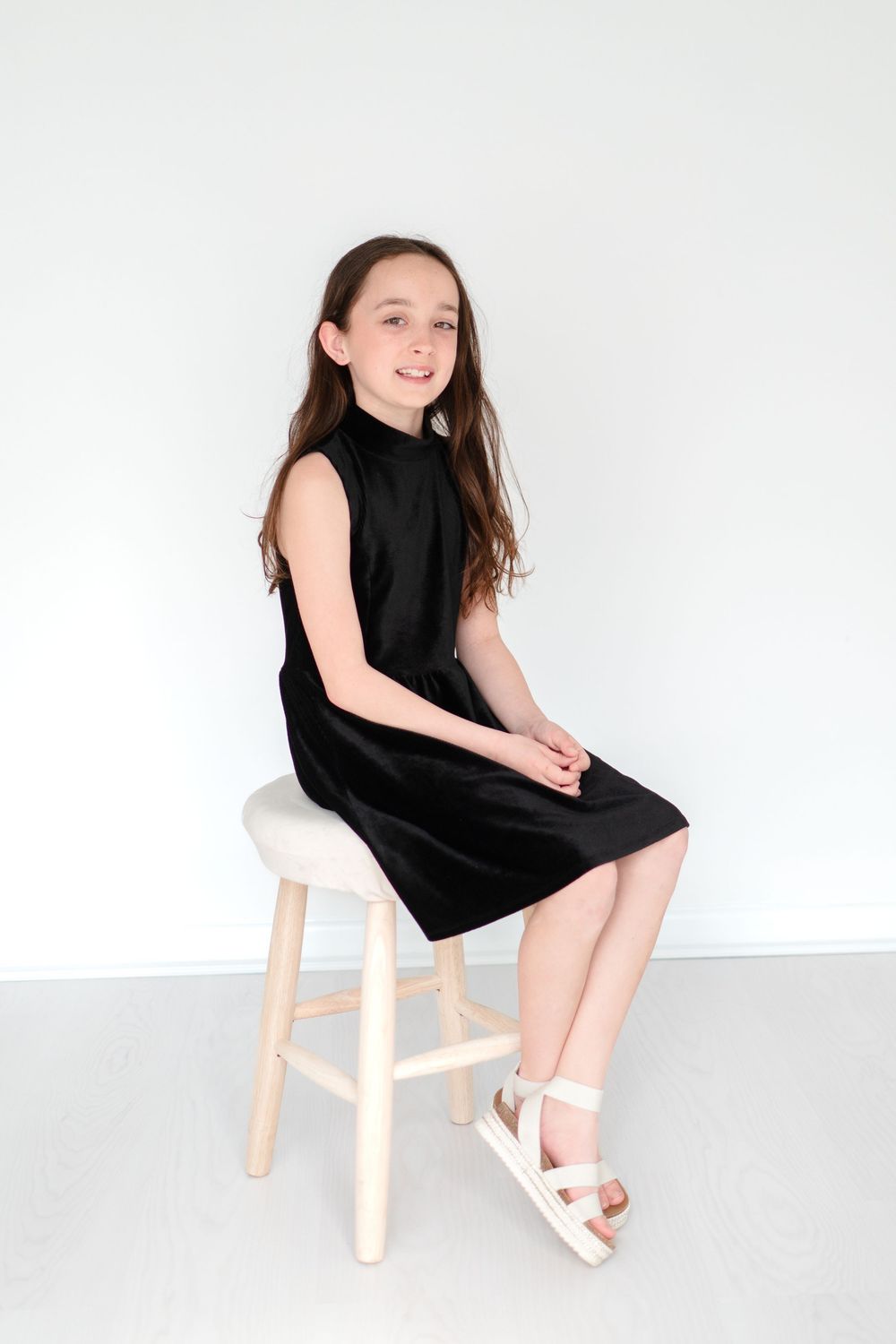 A young figure in a sleeveless black dress sits casually on a wooden stool against a white background.