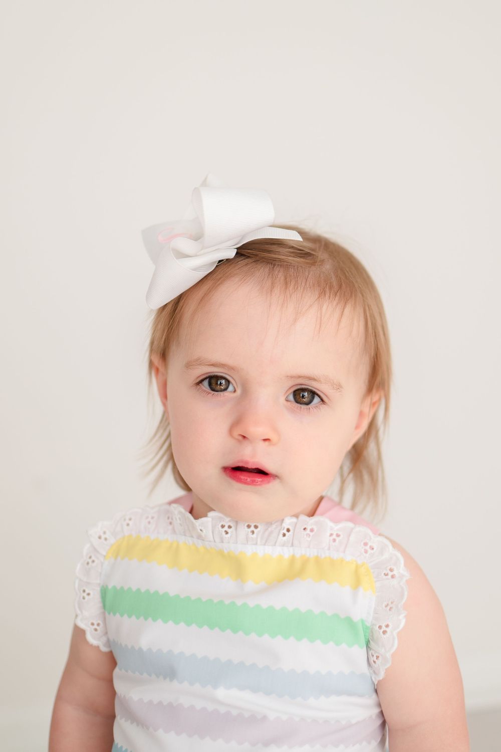 A toddler wearing a white bow and pastel striped dress poses against a light background.