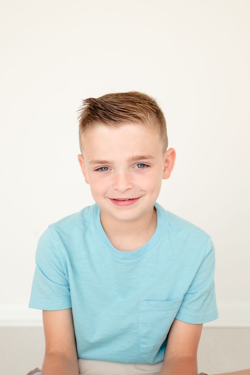 Child wearing light turquoise t-shirt sits against white background in casual portrait pose.