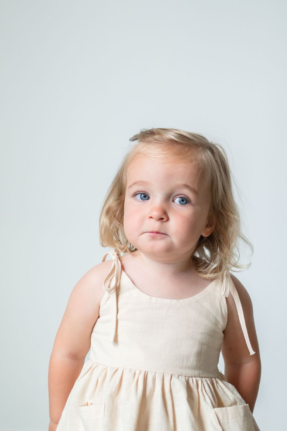 Portrait against white background wearing a cream colored dress with tied shoulders.