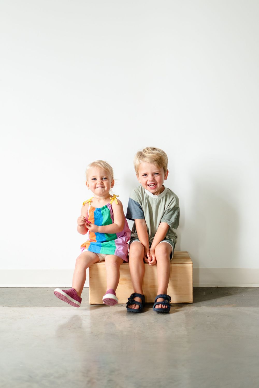 Two children sitting together on a wooden bench against a white wall.
