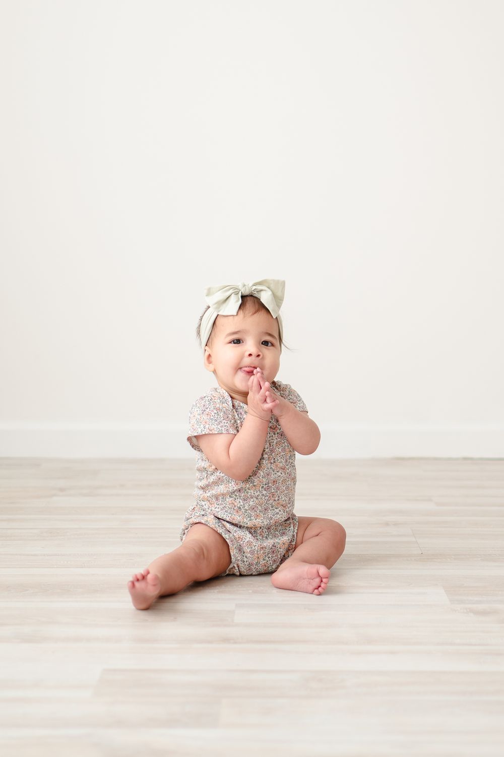 Baby wearing floral outfit and bow headband sitting on wooden floor against white wall.