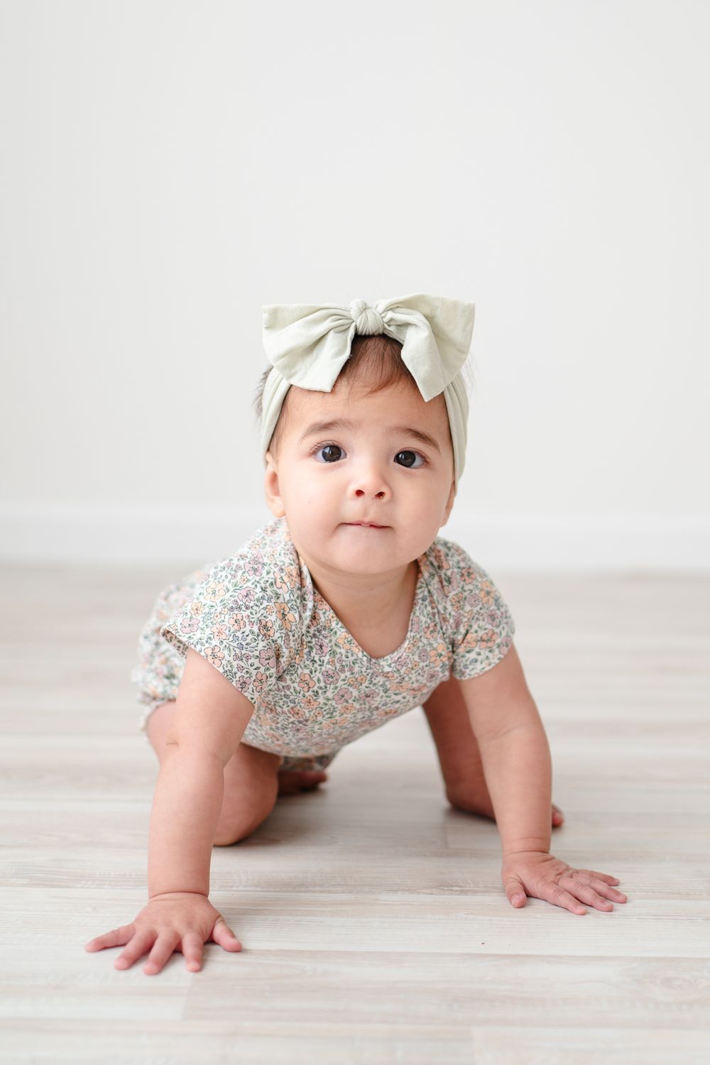 Baby in floral romper and white bow headband crawling on wooden floor.