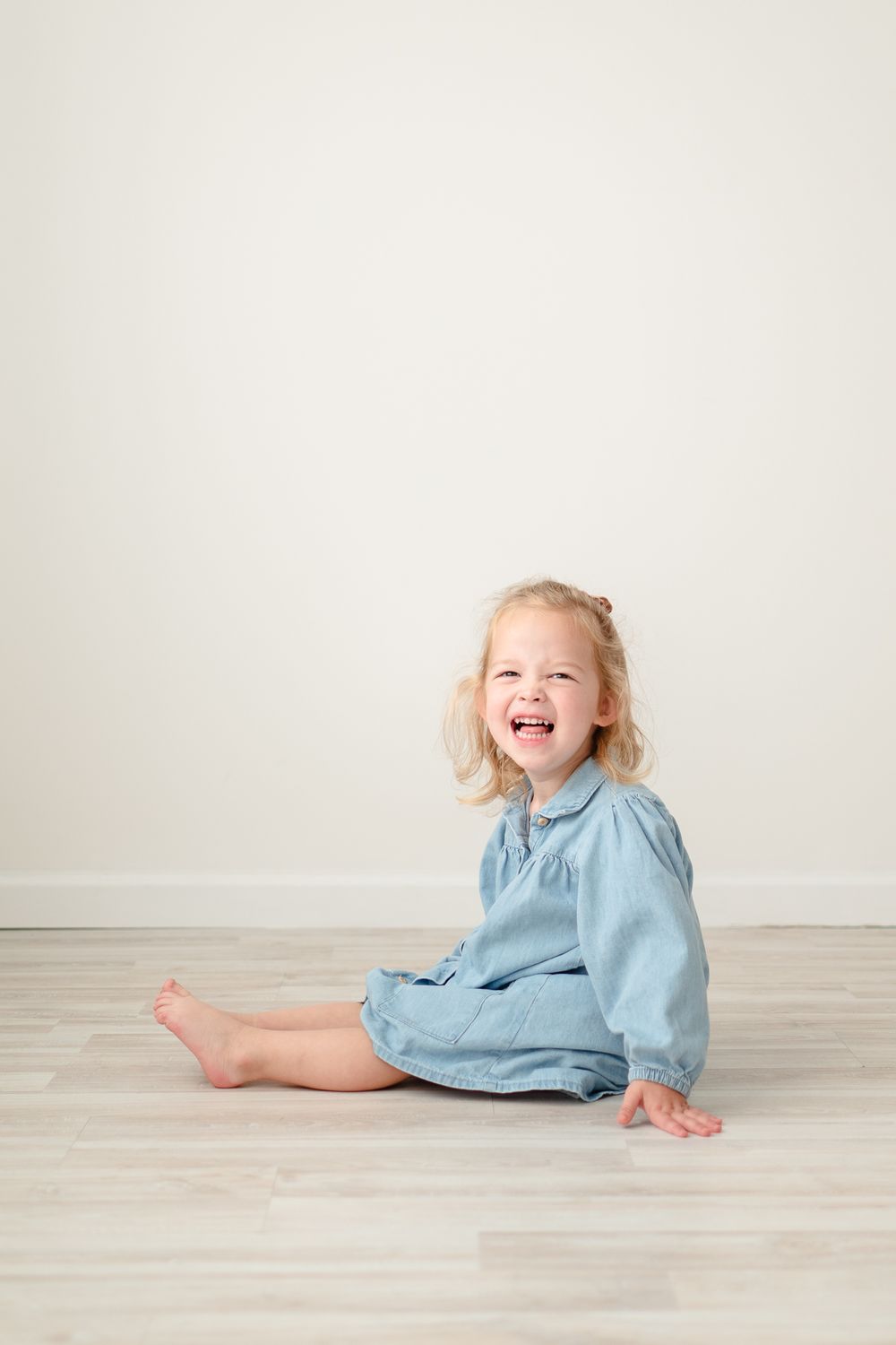 Child in denim dress sitting and laughing on wooden floor against white wall.