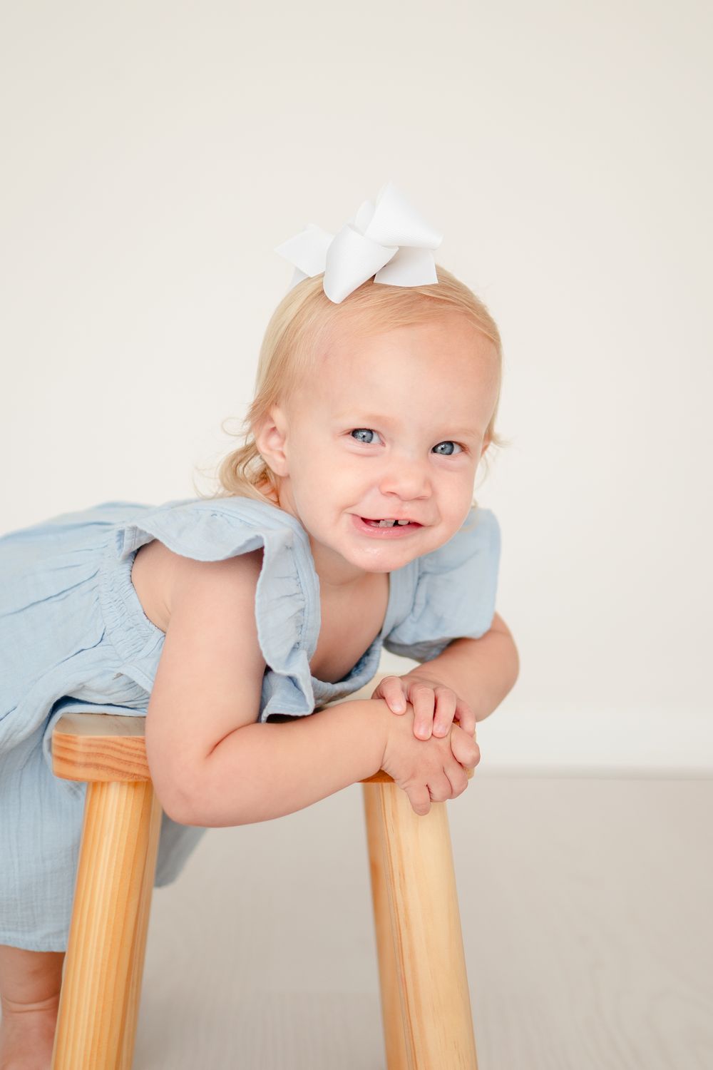 Toddler in light blue ruffled dress with white bow accessory leans playfully on wooden chair against neutral backdrop.