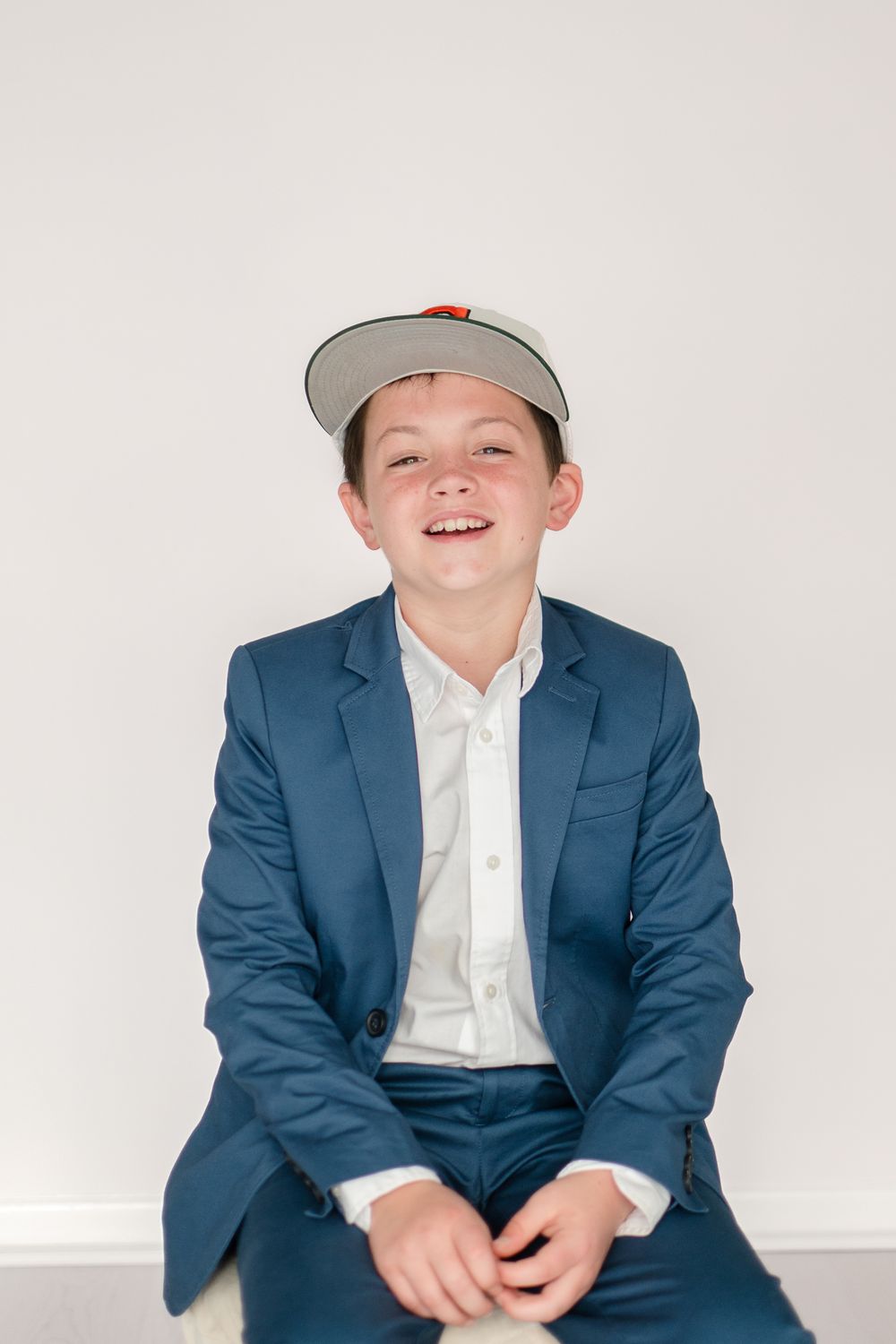 A person in a navy blue suit and baseball cap sits smiling against a white wall.