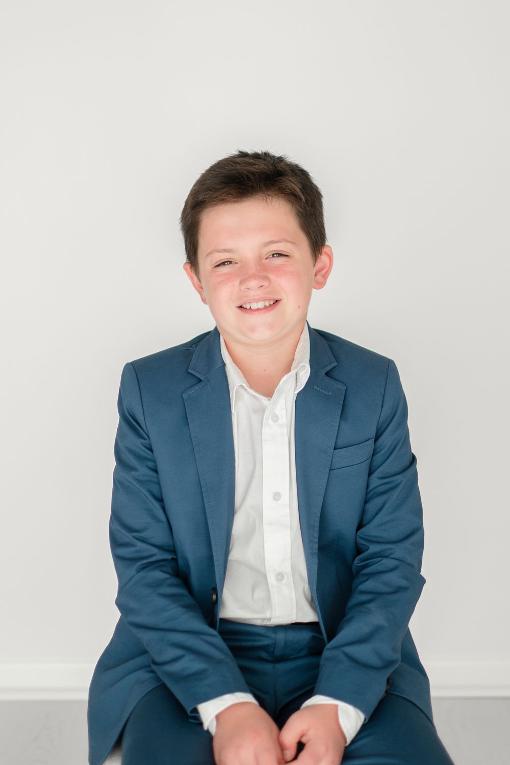 Young child in a smart blue suit jacket and white dress shirt sits and smiles against a light gray background.